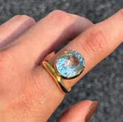 A gold ring with a round light blue gemstone worn on a finger, shown in a close-up shot against a dark textured background.